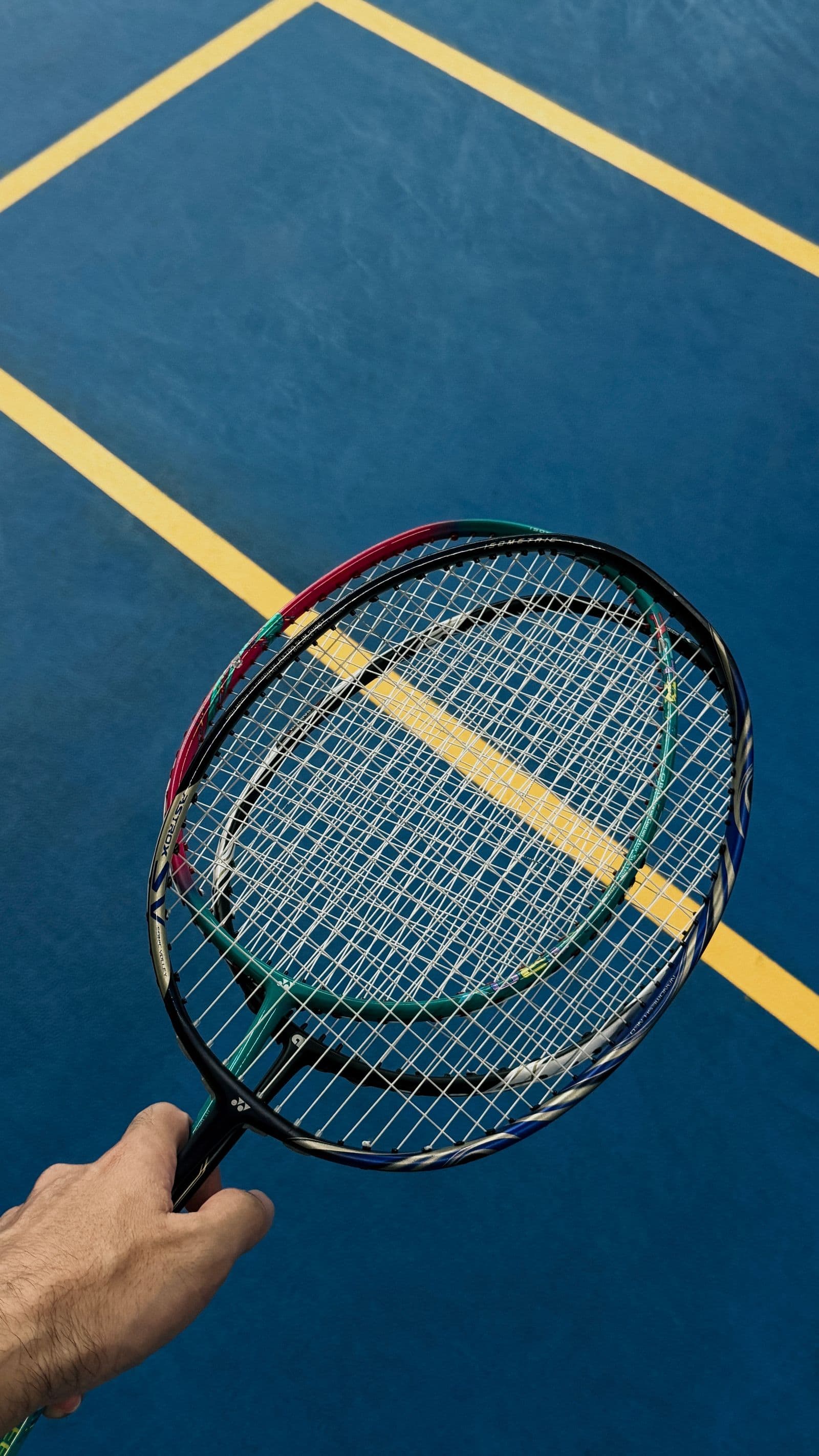 A hand holding a badminton racket overhead on a blue indoor court with yellow boundary lines