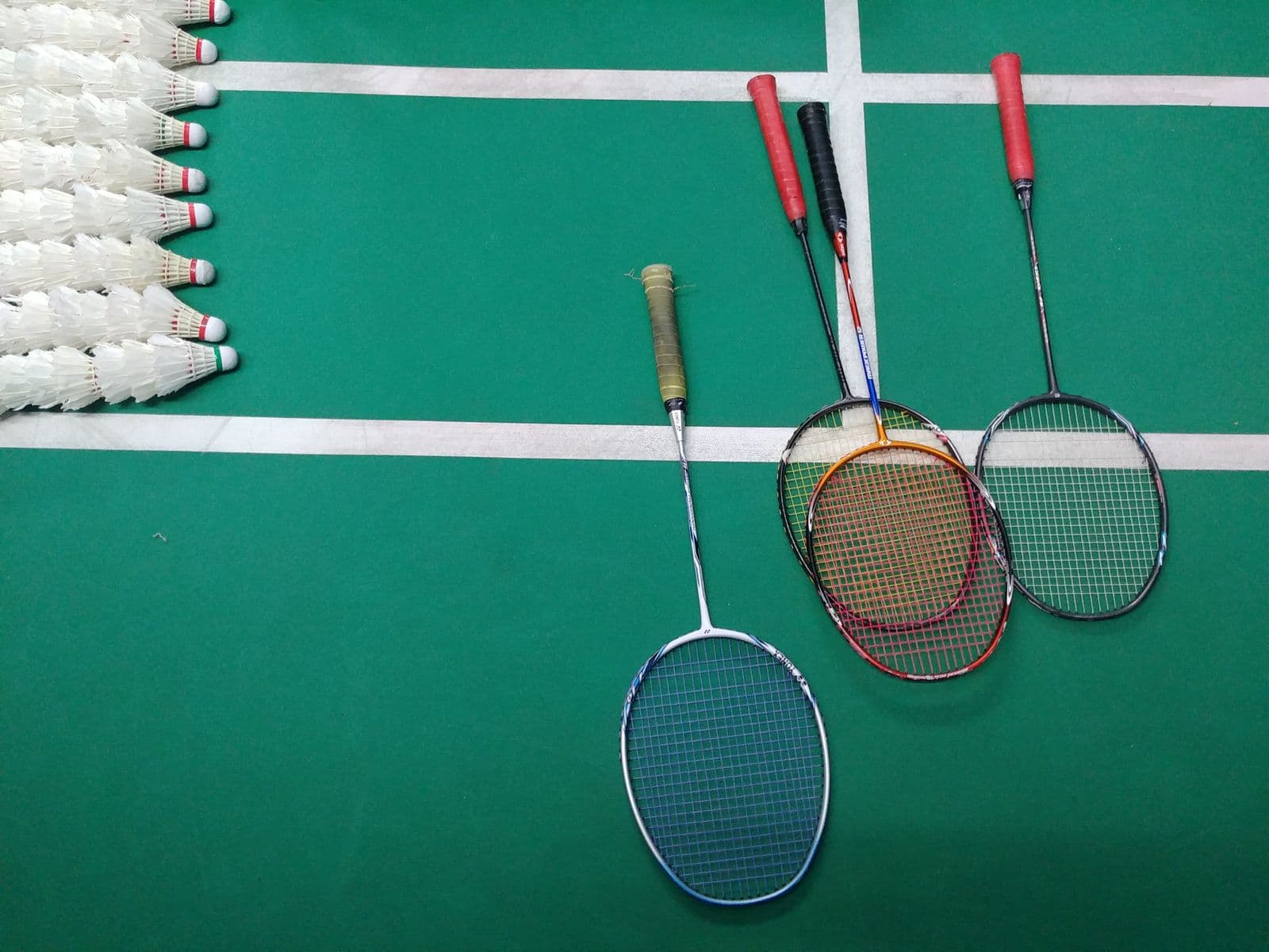 Four different badminton rackets and a stack of shuttlecocks arranged on a green indoor court