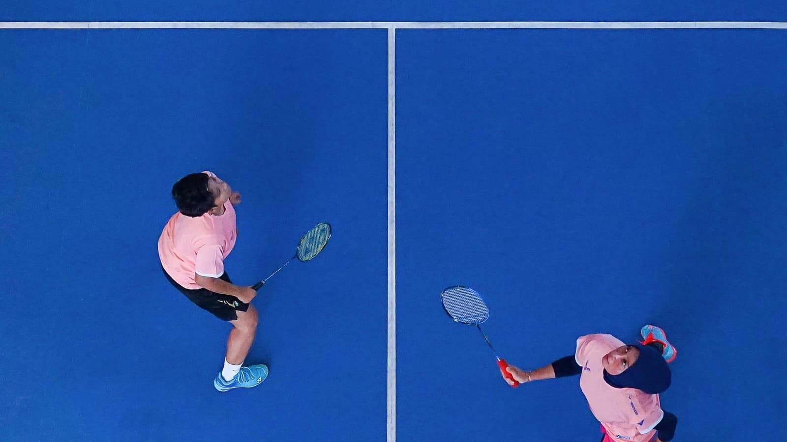 Aerial view of two badminton doubles players on a blue indoor court with court markings visible