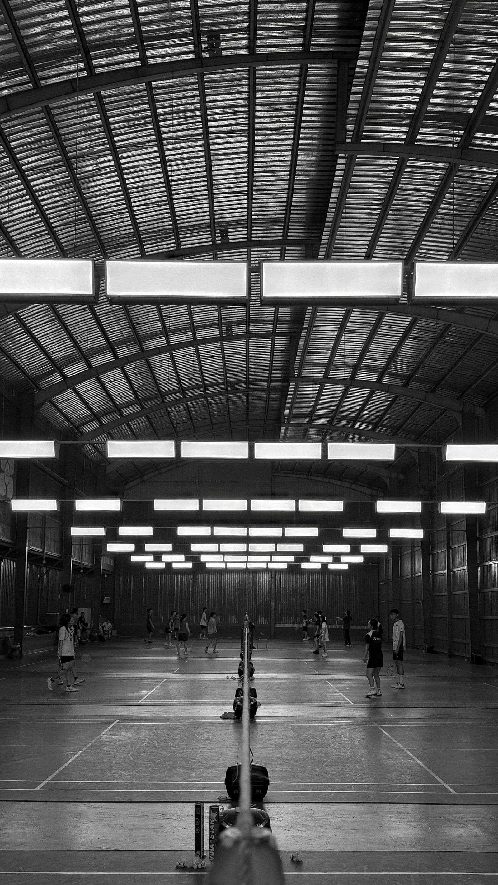 Monochrome wide-angle view of an indoor badminton hall from behind the net, with distant players and an arched industrial roof