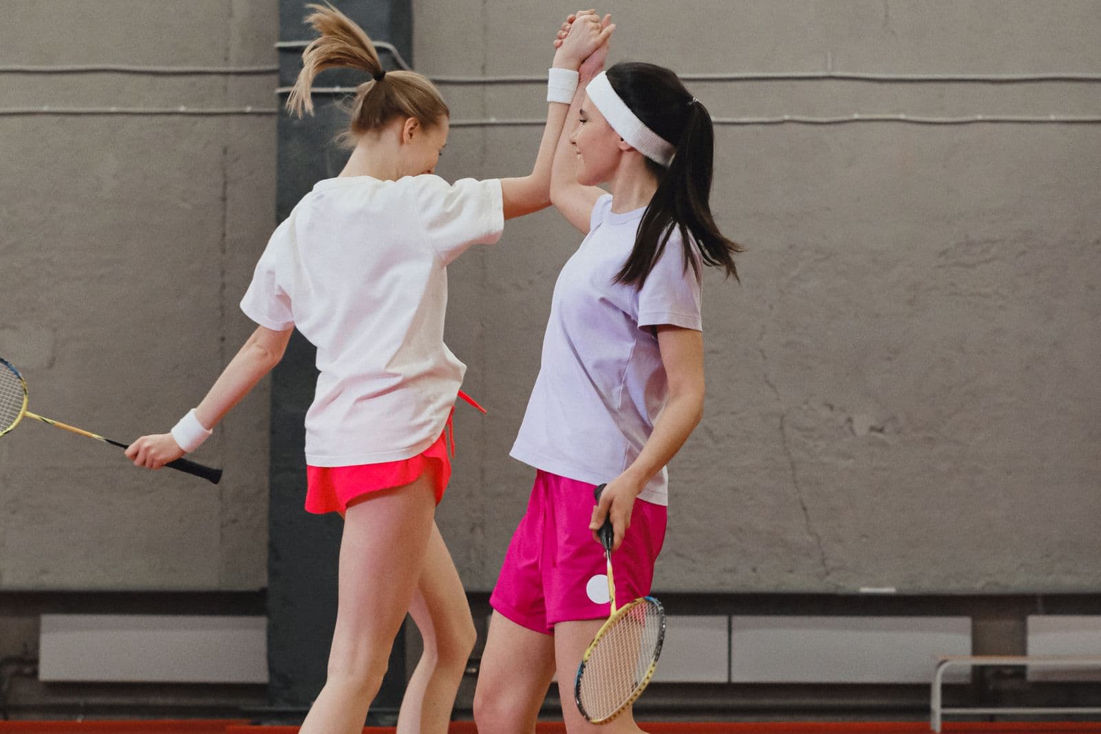 Two badminton doubles partners high-fiving mid-court in an indoor sports hall, rackets in hand