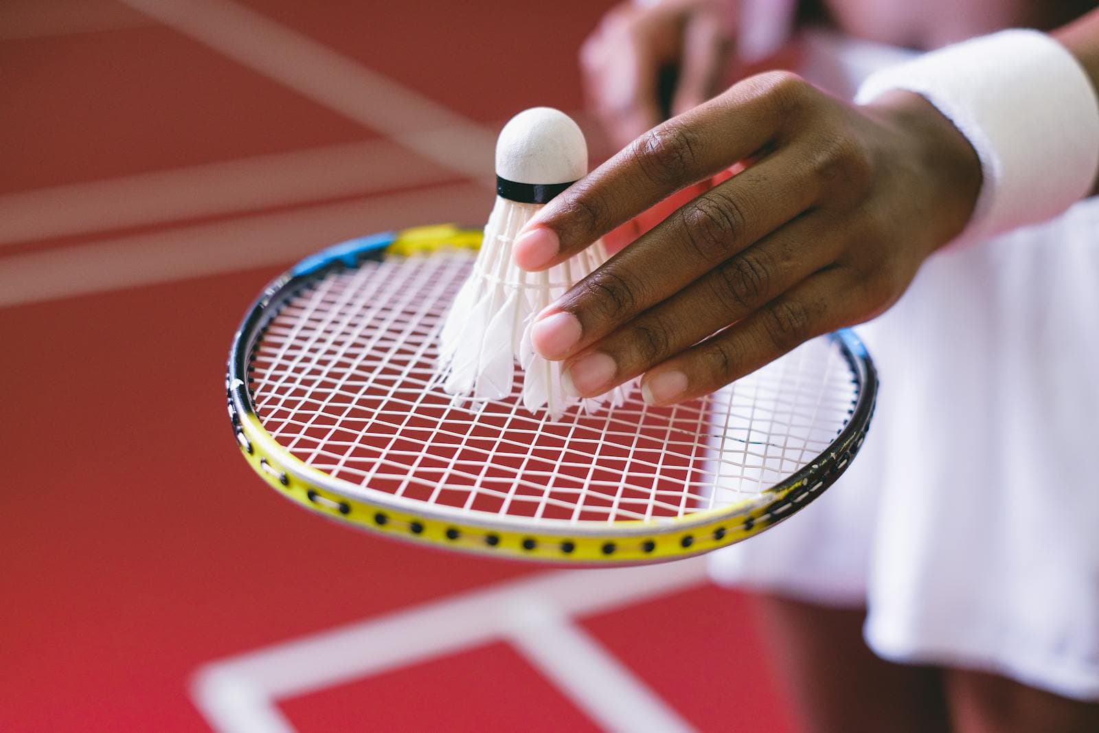 Close-up of a hand holding a shuttlecock above the strings of a badminton racket, about to serve, on a red indoor court