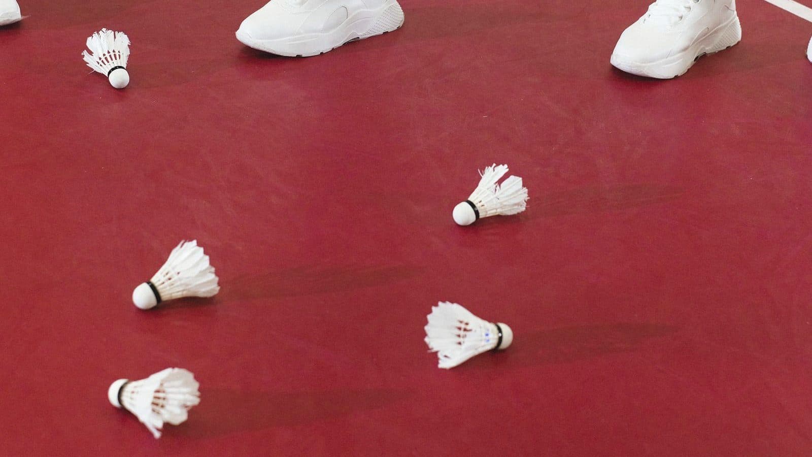 Feather shuttlecocks scattered across a red indoor badminton court with players' shoes at the edge of the frame