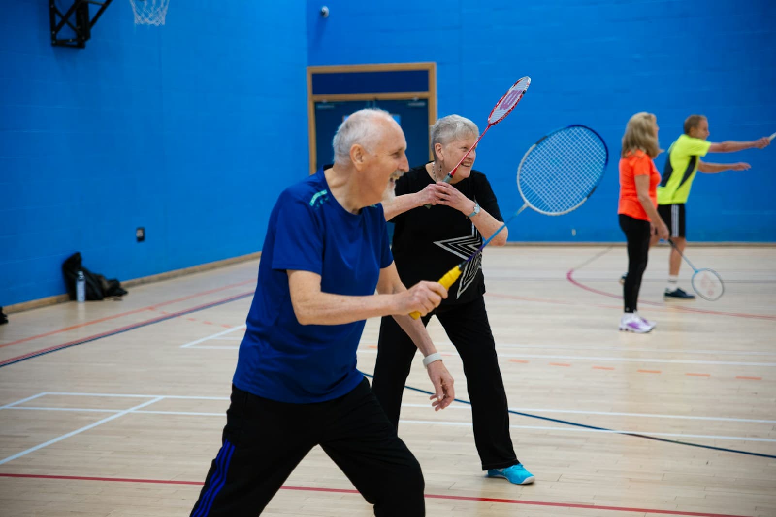 Adults playing doubles badminton on an indoor club court, front and back players in action