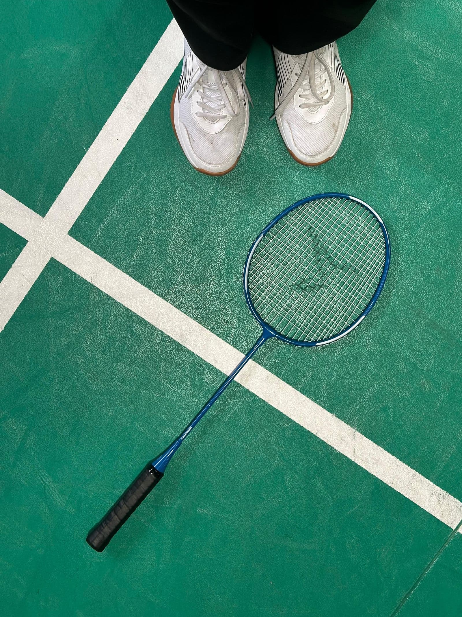 Top-down view of a badminton racket and a pair of white court shoes on a green indoor court with white line markings