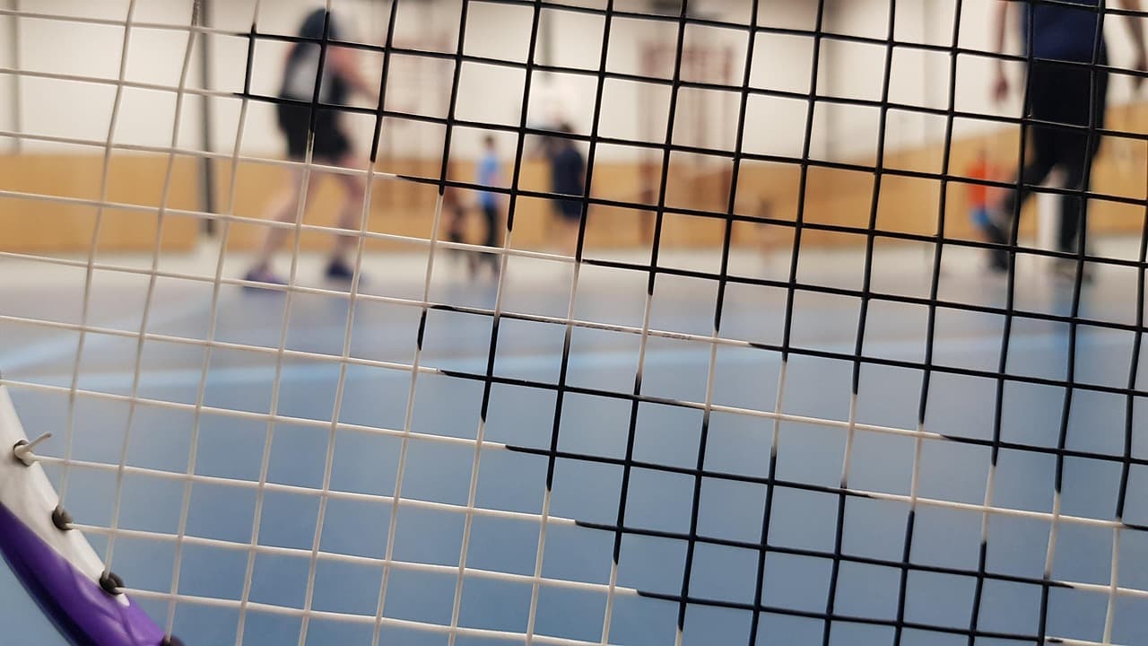 A blue indoor badminton court seen through the net and racket strings, with club players standing between rallies in the background