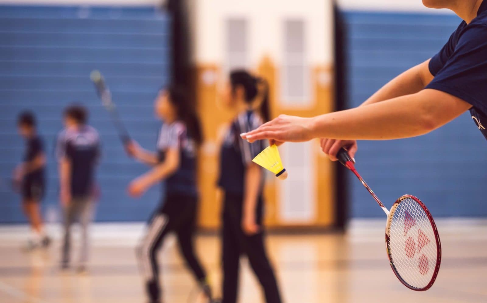 A player's hand holding a shuttlecock and racket before serving, with blurred club players moving in the background of a wooden sports hall
