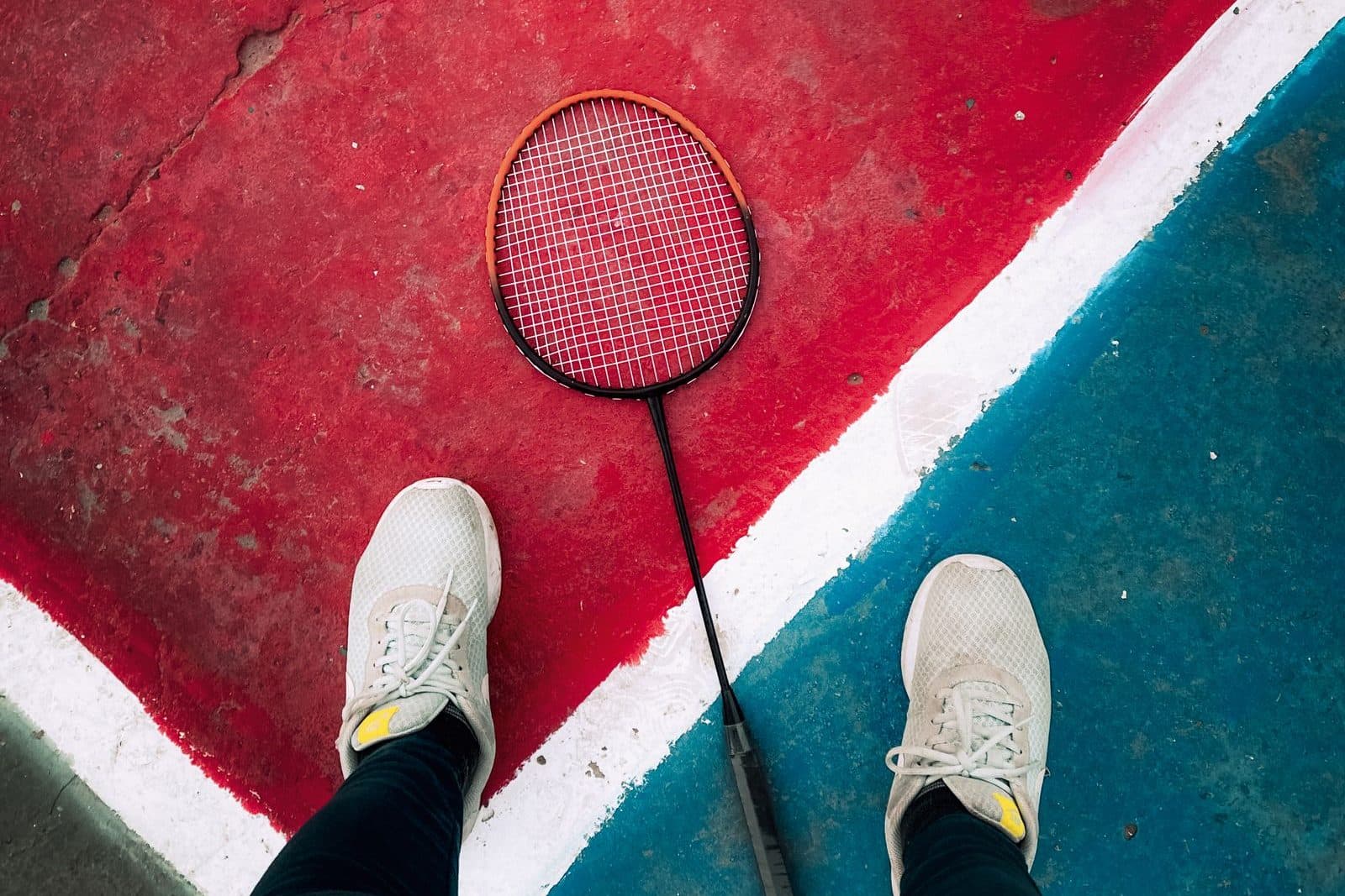 Top-down view of a player's shoes on either side of a badminton racket lying across the painted lines of a red and blue court