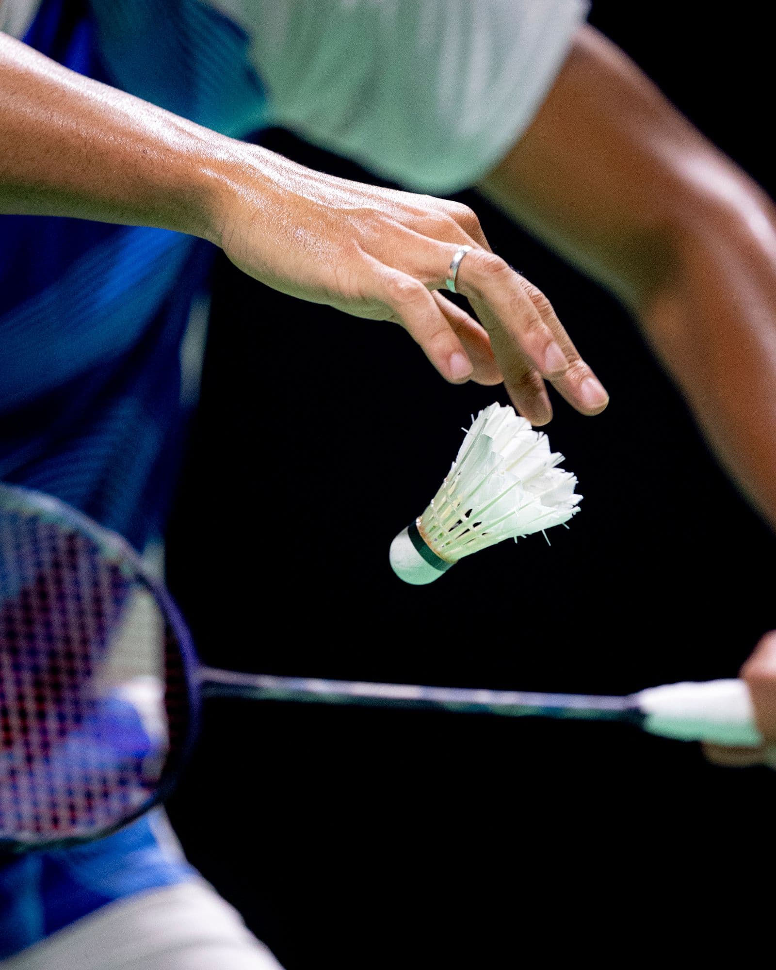 Close-up of a player's hand releasing a shuttlecock above the racket head at the moment of serve, against a dark indoor background