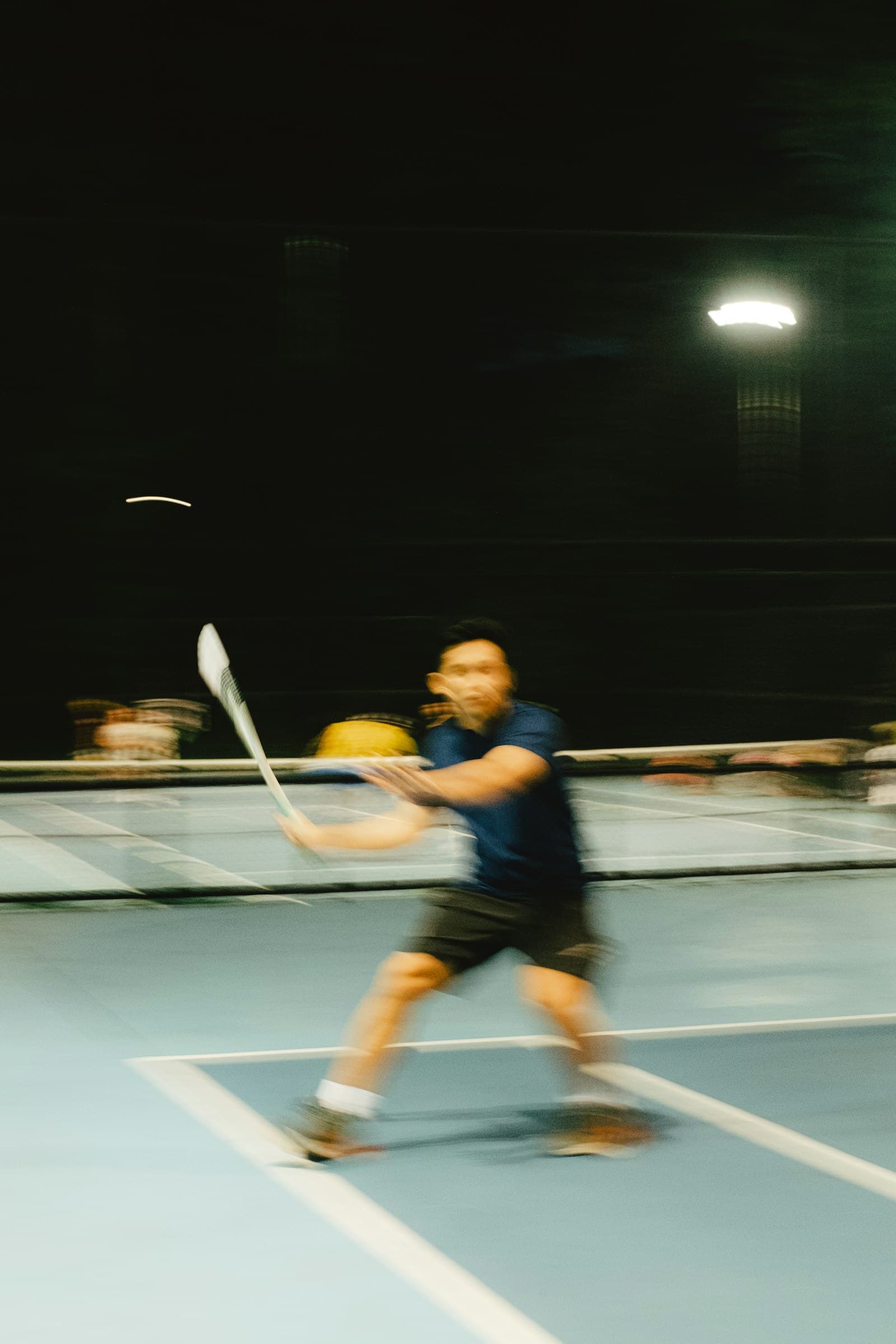Motion-blurred badminton player mid-swing on an indoor court under a single dramatic overhead light at night