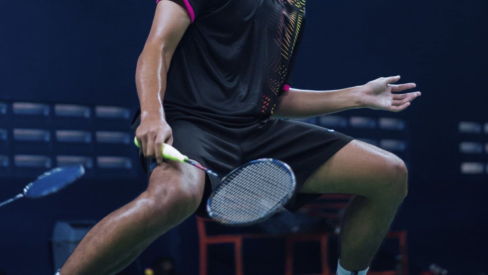 A doubles player low and wide in a defensive stance with the racket held out in front, on a dark indoor court