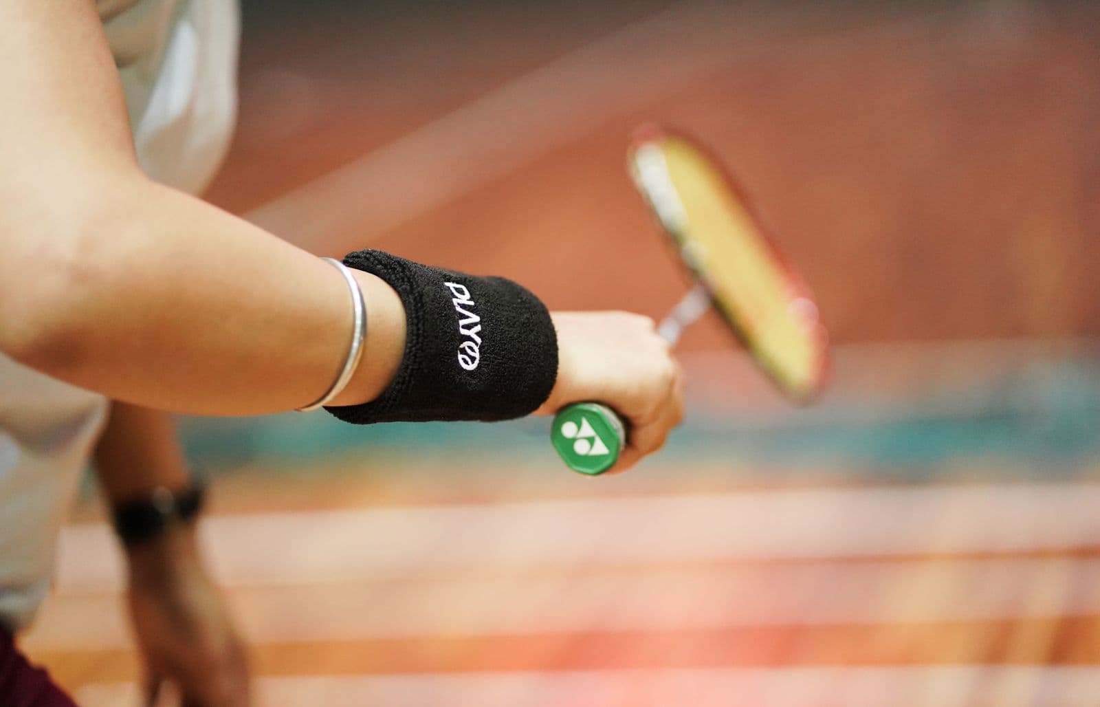 Close-up of a player's forearm and hand gripping a badminton racket handle, with a warm-toned indoor court blurred behind