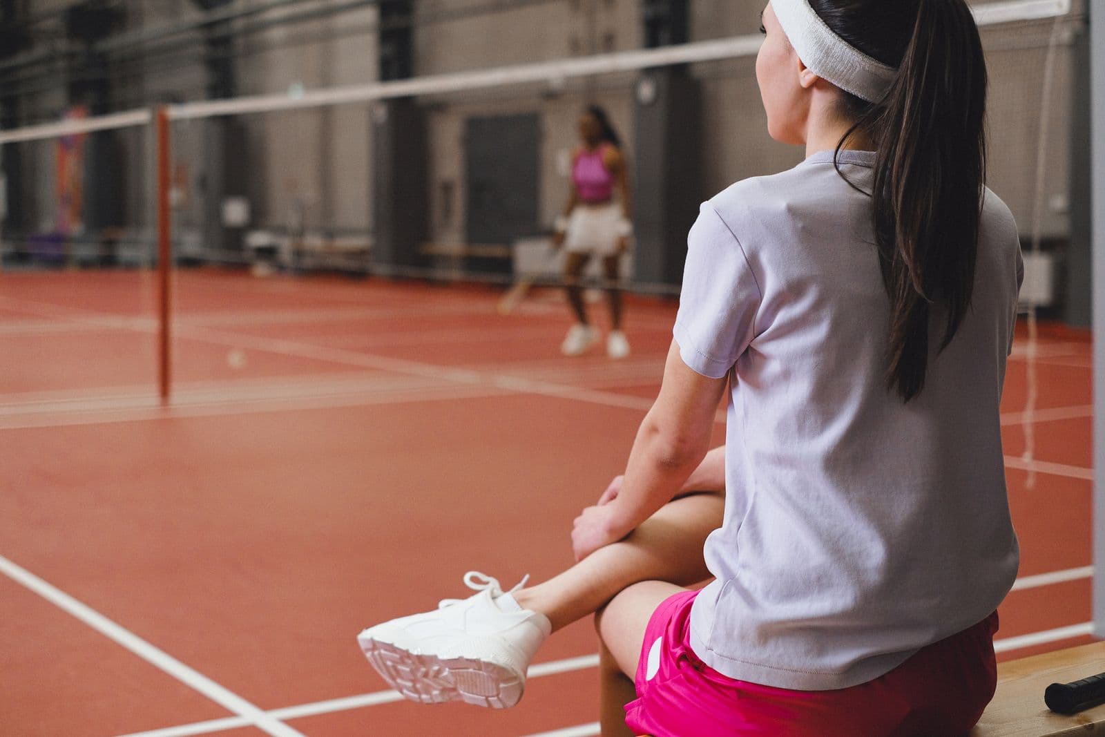 A player sitting courtside with her back to the camera, watching a rally on a red indoor badminton court
