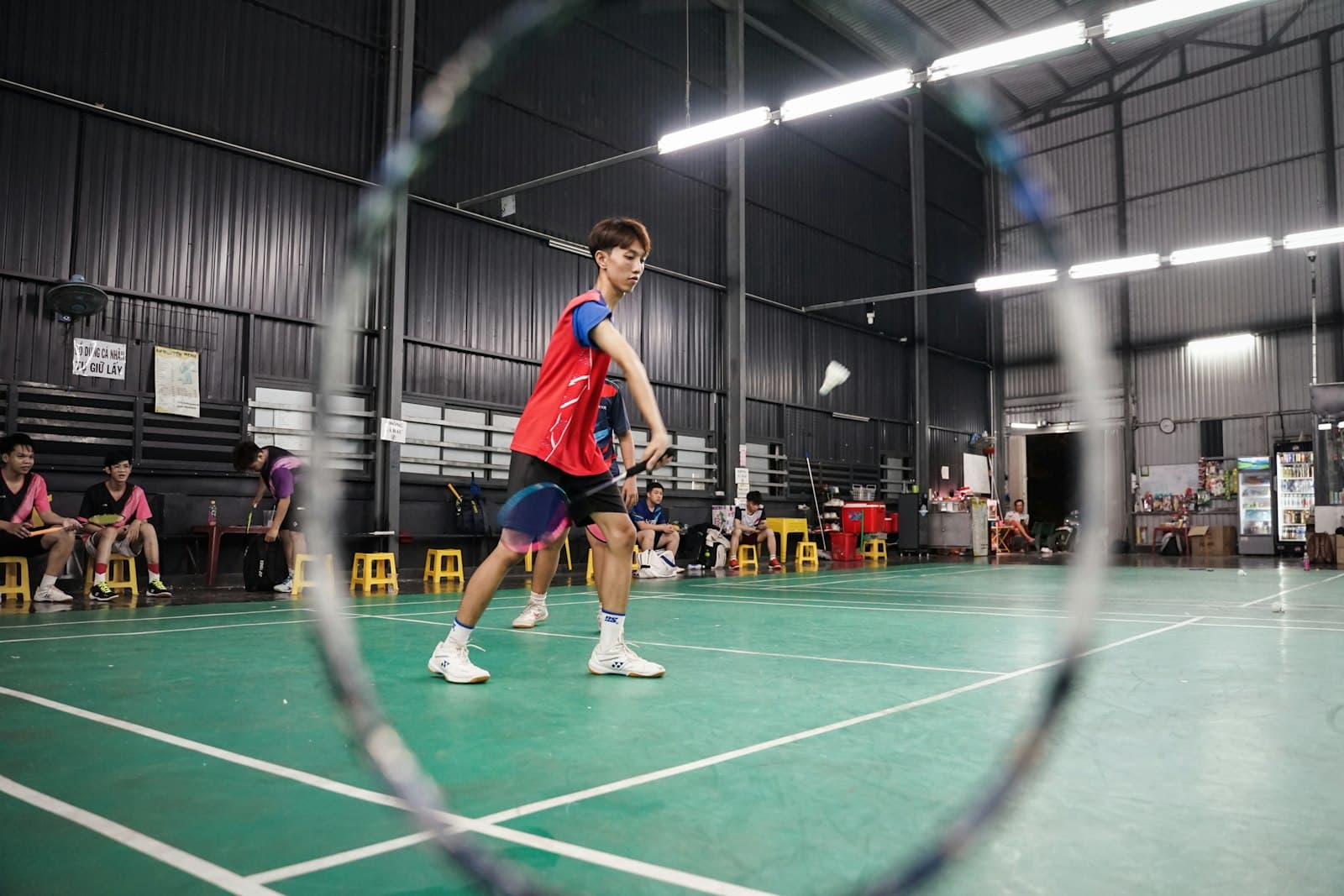 A badminton club court seen through the head of a racket, with a player mid-rally on a green indoor floor and others waiting courtside