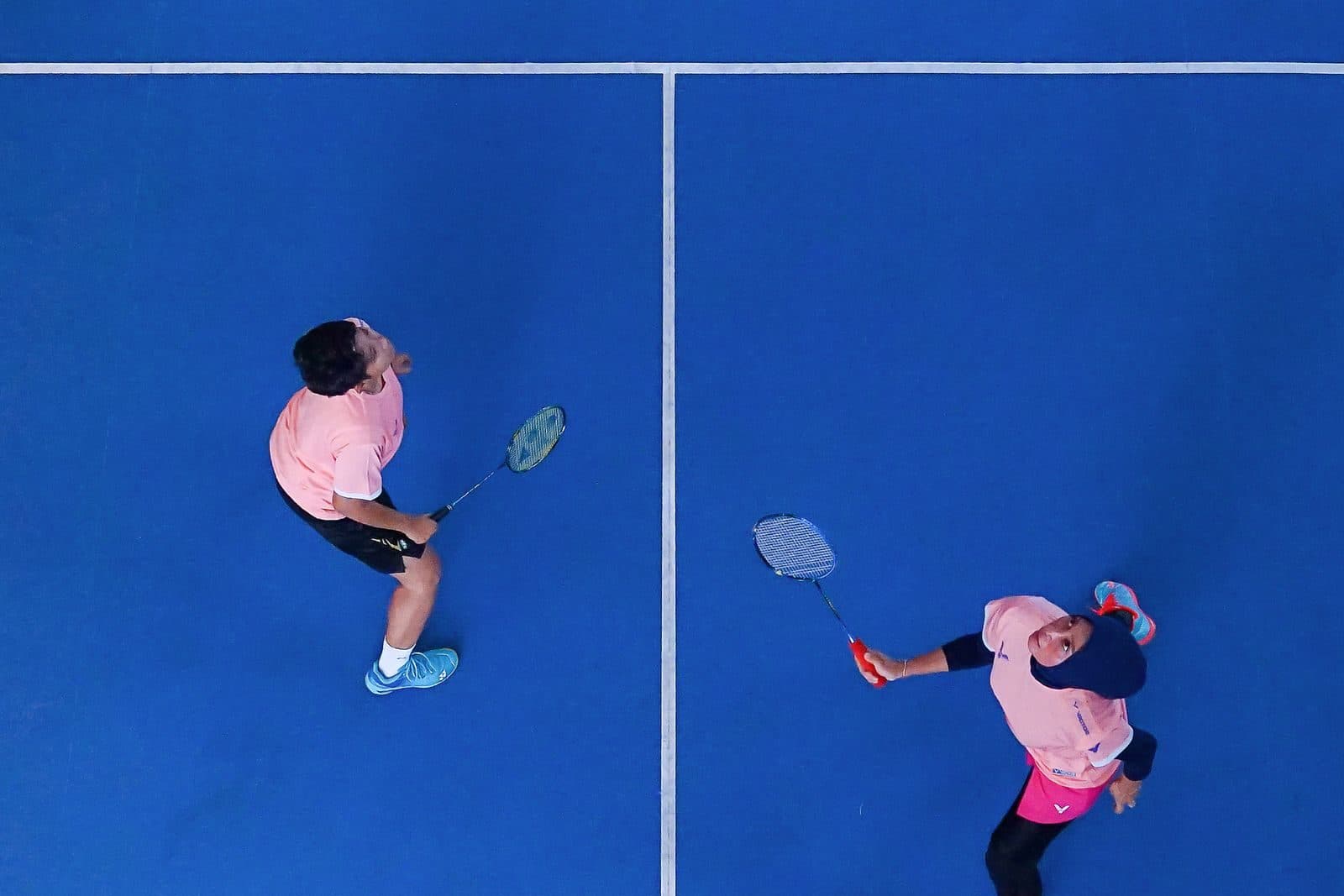 Overhead view of two badminton doubles partners on either side of the centre line on a blue indoor court