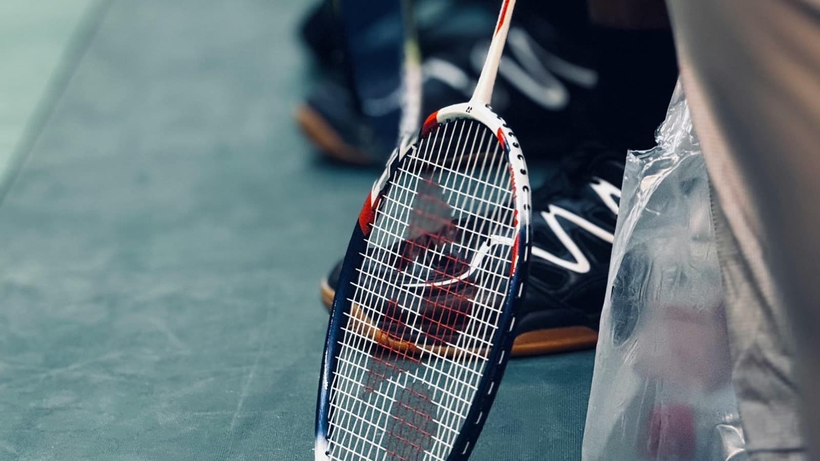 Close-up of a badminton racket resting on an indoor sports court floor with players in soft focus in the background