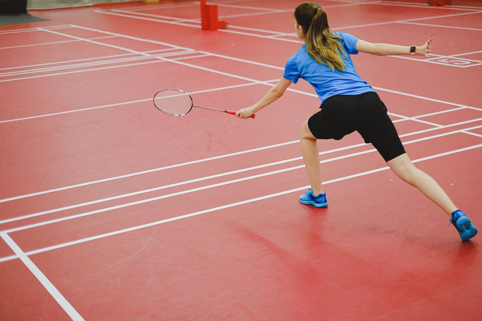 Back view of a player mid-rally on a red indoor badminton court, racket raised toward the far side