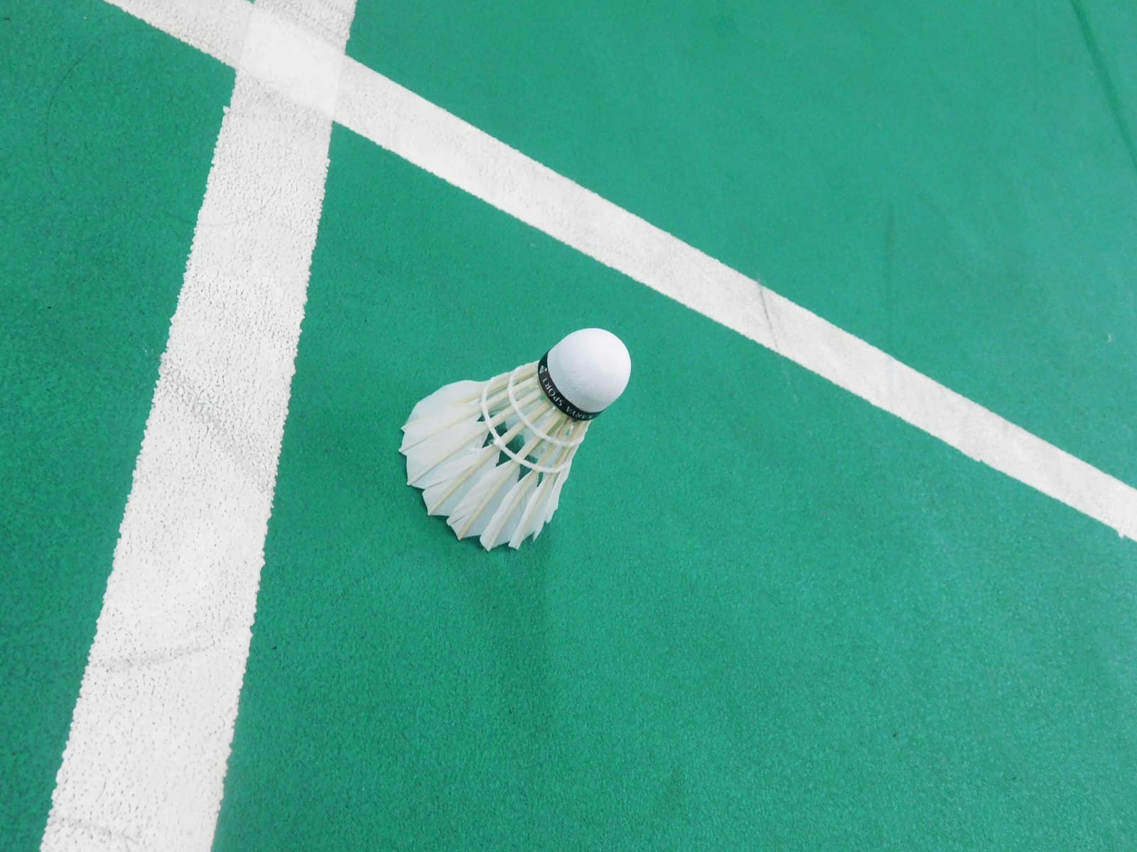 A white shuttlecock standing upright on the painted lines of a green badminton court between rallies