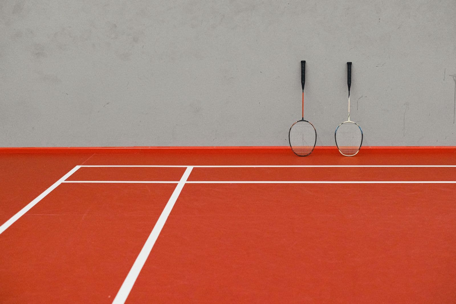 Two badminton rackets leaning against a wall on an indoor red court