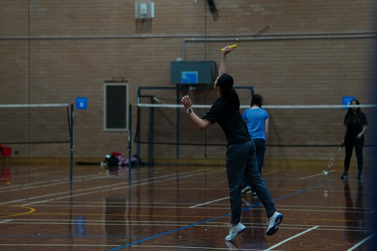 A club player reaching for an overhead in a wooden-floored sports hall with a brick wall, other players blurred in the background