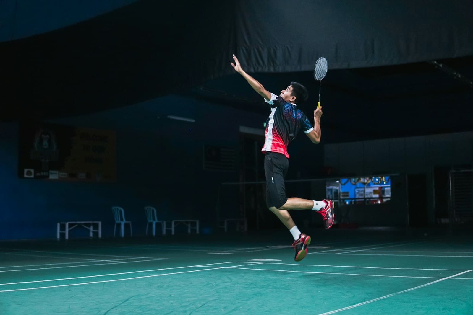 A player mid-air in a jump smash on a dimly lit green indoor badminton court, seen from the side