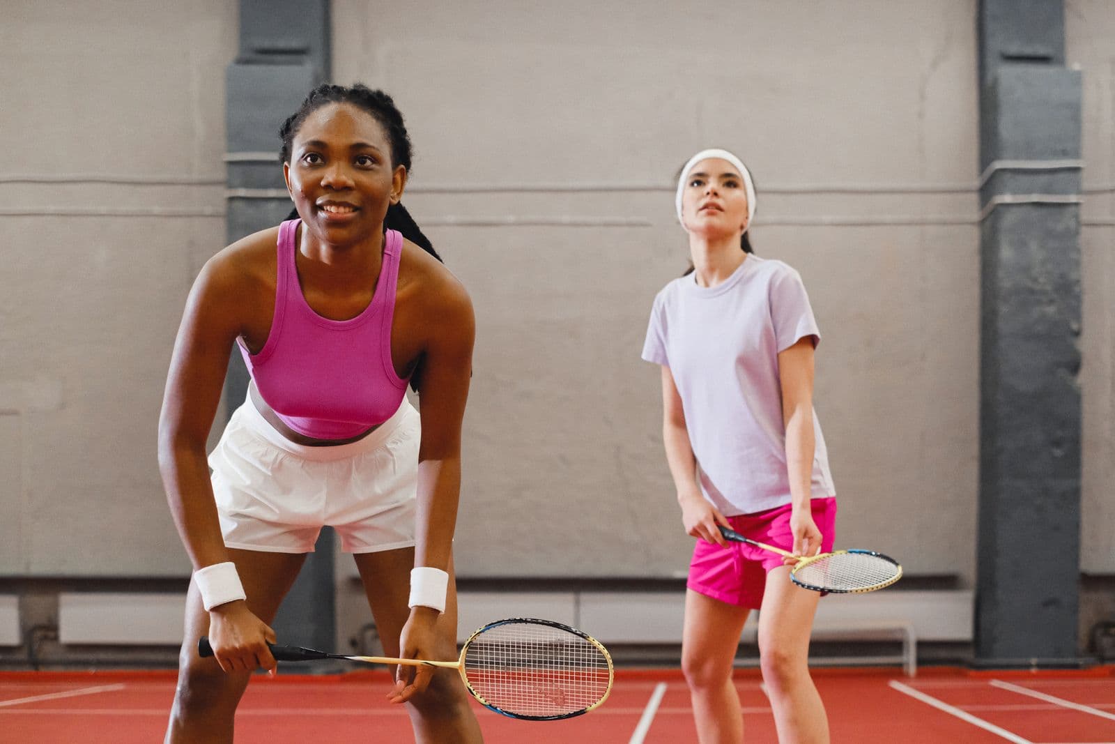 Two women playing doubles badminton on an indoor court, mid-rally with rackets raised